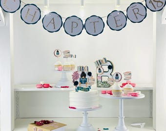 May include: A white cake with a book-themed topper that says "Chapter 18 Bella". The cake is on a white cake stand. There are cupcakes with book-themed toppers on a white cupcake stand. There is a banner that says "Chapter 18" in blue and gray with gold glitter. There is a book-shaped box that says "Bella" in gold glitter.