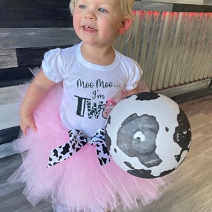 May include: A young child wearing a white shirt with the text "Moo Moo... I'm Two" and a pink tutu with a black and white cow print bow. The child is holding a black and white cow print balloon.