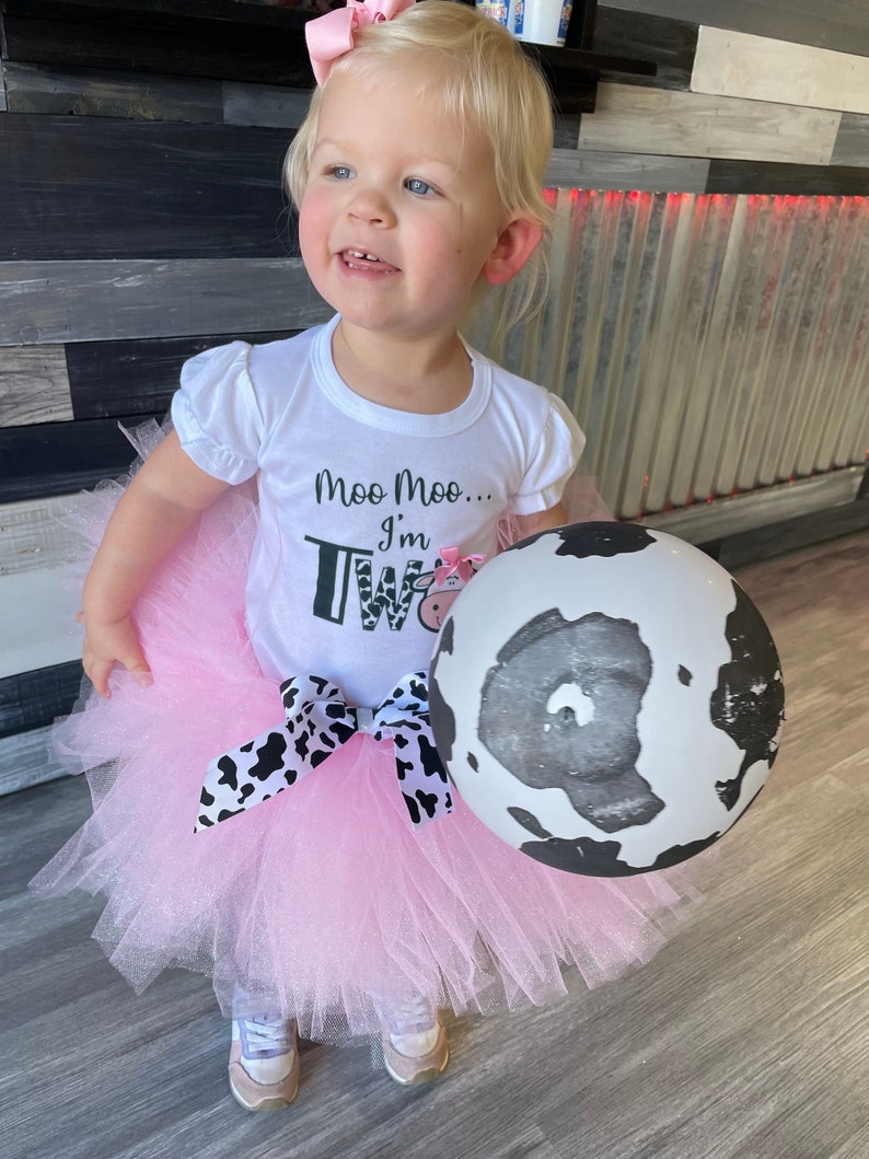 May include: A young child wearing a white shirt with the text "Moo Moo... I'm Two" and a pink tutu with a black and white cow print bow. The child is holding a black and white cow print balloon.