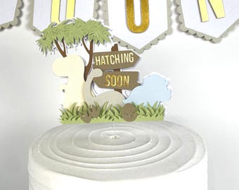 May include: A white cake with a dinosaur-themed topper and the words "Hatching Soon." A banner above spells out "OON" in gold letters. The cake has a textured, swirled frosting design. The topper includes a tree and grass.