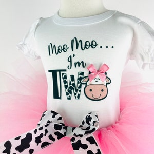 May include: A pink tutu with a black and white cow print bow. A white t-shirt with the text "Moo Moo... I'm Two" and a cow graphic.
