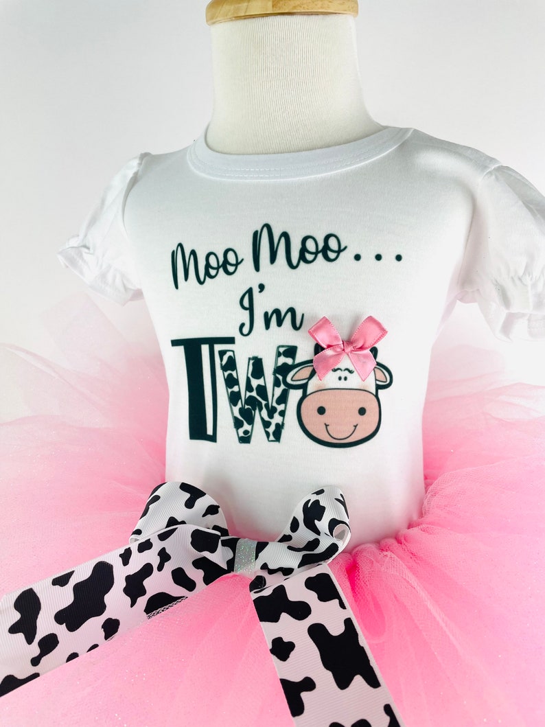 May include: A pink tutu with a black and white cow print bow. A white t-shirt with the text "Moo Moo... I'm Two" and a cow graphic.