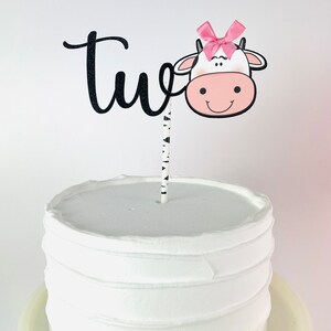 May include: A white cake with black and white striped frosting and a black and white cow cake topper with a pink bow. The topper says "two" in black cursive.