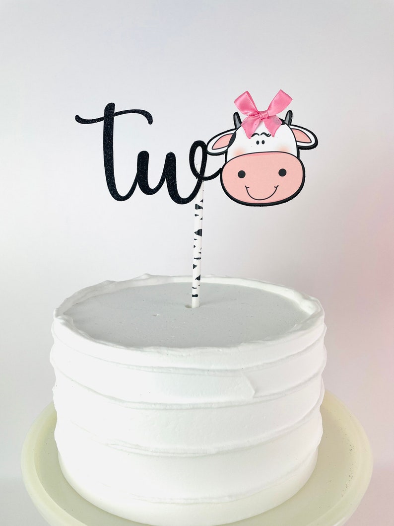 May include: A white cake with black and white striped frosting and a black and white cow cake topper with a pink bow. The topper says "two" in black cursive.
