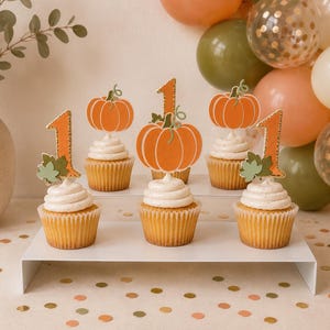 Puede incluir: Cinco cupcakes con glaseado blanco dispuestos en un soporte blanco de varios niveles. Cada cupcake presenta un adorno de papel decorativo, incluyendo calabazas naranjas y el número uno, con pequeñas hojas verdes y bordes dorados brillantes.