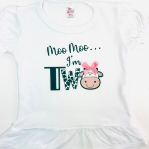 May include: White short-sleeved shirt with a black and white cow print 'TWO' and the text 'Moo Moo... I'm Two' with a pink bow on the cow's head.