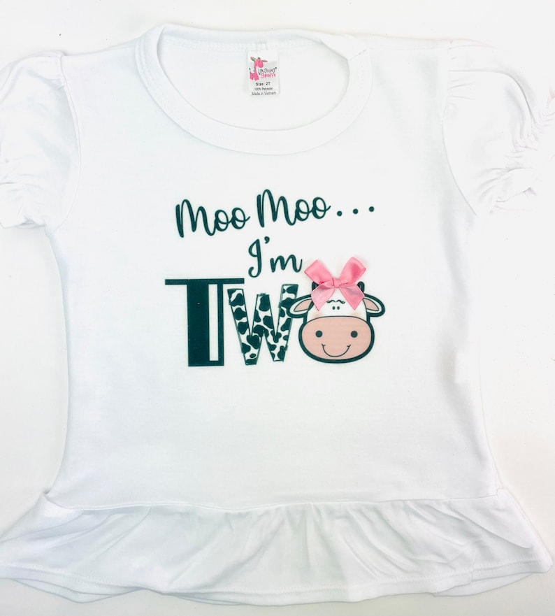May include: White short-sleeved shirt with a black and white cow print 'TWO' and the text 'Moo Moo... I'm Two' with a pink bow on the cow's head.