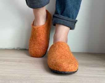 May include: A pair of orange felt slippers with black soles. The slippers are worn by a person wearing blue jeans.