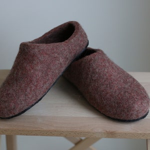 Könnte beinhalten: Ein Paar braune Wollhausschuhe auf einem Holzhocker. Die Hausschuhe sind aus einem weichen, flauschigen Material gefertigt und haben ein schlichtes, klassisches Design.