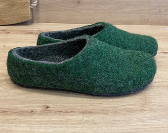 Könnte beinhalten: Ein Paar grüne Filzschuhe mit dunkelgrauem Innenfutter und dunkelblauer Sohle. Die Hausschuhe bestehen aus einem strukturierten, wollähnlichen Material. Der Hintergrund ist eine hellbraune Holzoberfläche.