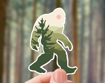 May include: A sticker of a Bigfoot silhouette with a forest scene inside. The sticker is being held by a hand in front of a blurred background of trees.