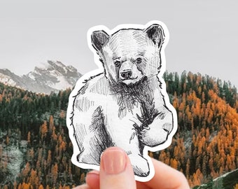 May include: A black and white line drawing of a bear cub, printed on a sticker. The sticker is held by a hand against a backdrop of a mountain range with snow-capped peaks and a forest of pine trees.