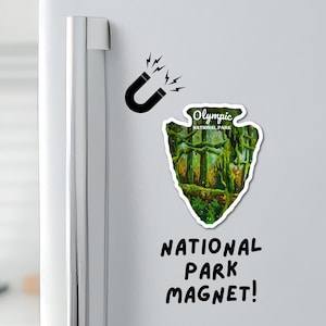 May include: A green and brown National Park magnet shaped like an arrowhead with the text "Olympic National Park" on it. The magnet is attached to a white refrigerator with a black horseshoe magnet and the text "NATIONAL PARK MAGNET!" written below it.