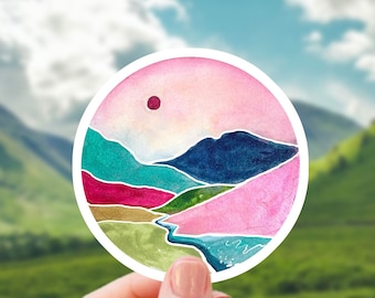 May include: A round sticker with a watercolor painting of a mountain range with a pink, blue, green, and gold color scheme. The sticker has a white border and a red sun in the upper right corner.
