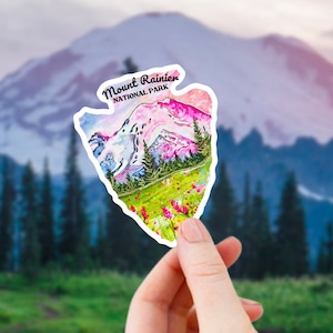 May include: A sticker featuring a watercolor illustration of Mount Rainier National Park. The sticker is shaped like a mountain peak and has the text "Mount Rainier National Park" at the top.