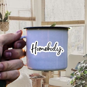 Pu&ograve; includere: Una tazza di smalto bianca con un bordo nero e un adesivo che dice "Homebody". La tazza &egrave; tenuta da una mano in un maglione verde.