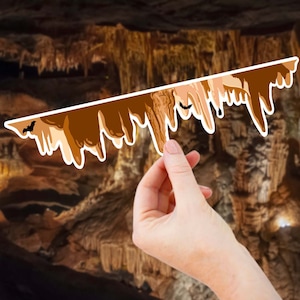 May include: A hand holds up a brown and tan sticker of cave formations, with a silhouette of a bat in the top left corner. The sticker is against a blurred background of a cave with brown and tan rock formations.