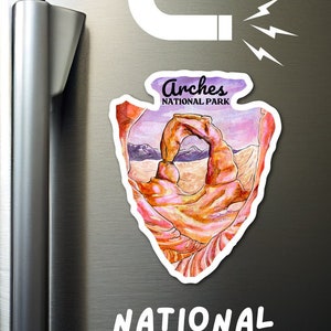 May include: A refrigerator magnet featuring a watercolor illustration of Delicate Arch in Arches National Park, Utah. The magnet is shaped like a pointed arrow and has the text "Arches National Park" printed on it. The text "NATIONAL PARK MAGNET!" is written below the magnet.