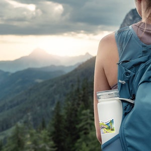 May include: A person wearing a blue backpack with a white water bottle attached to the strap. The water bottle has a sticker on it that says "The Rocky Mountains". The person is standing in front of a mountain range with a cloudy sky.
