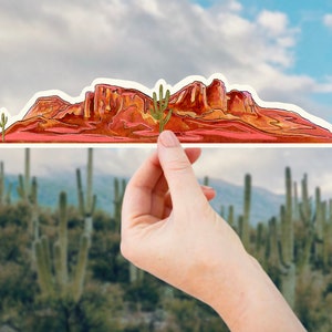 May include: A red and orange watercolour illustration of a mountain range with a green cactus in the foreground. The illustration is cut out and held in front of a blurred background of cacti.