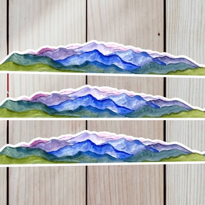 Pode incluir: Tr&ecirc;s adesivos de aquarela de cordilheiras com uma base verde e picos roxos, azuis e brancos. Os adesivos est&atilde;o sobre um fundo de madeira.