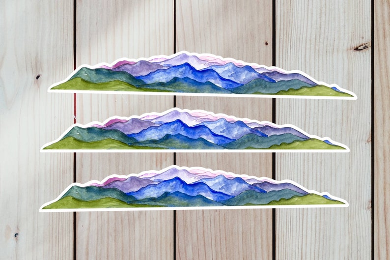Pode incluir: Tr&ecirc;s adesivos de aquarela de cordilheiras com uma base verde e picos roxos, azuis e brancos. Os adesivos est&atilde;o sobre um fundo de madeira.