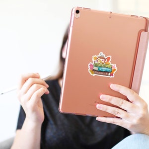 Op de afbeelding: Een ros&eacute;gouden iPad met een sticker van een stapel boeken, een theepot en herfstbladeren. De sticker heeft een aquarel stijl met tinten groen, blauw, oranje en bruin. De iPad wordt vastgehouden door een persoon die een zwart T-shirt draagt.