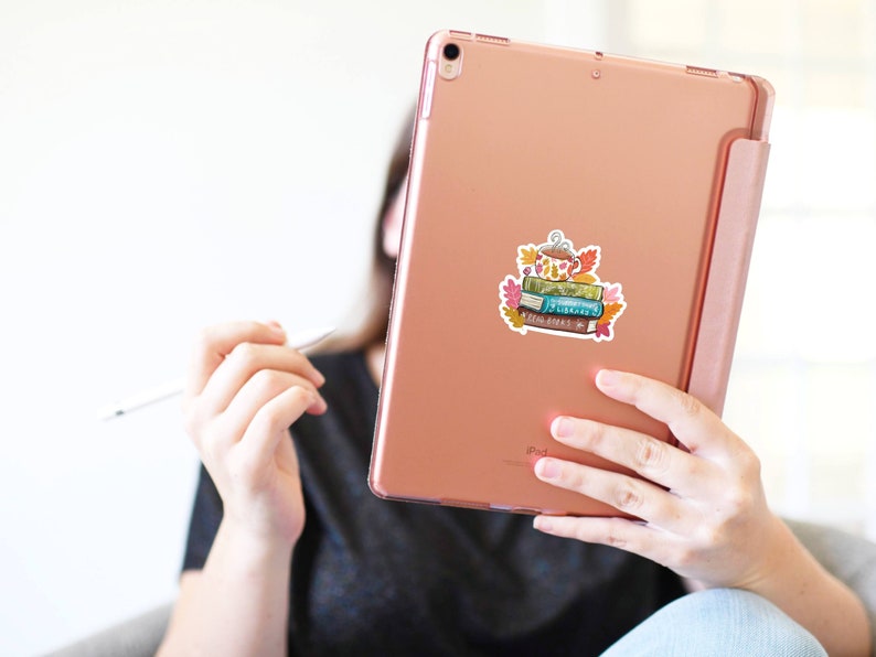 Op de afbeelding: Een ros&eacute;gouden iPad met een sticker van een stapel boeken, een theepot en herfstbladeren. De sticker heeft een aquarel stijl met tinten groen, blauw, oranje en bruin. De iPad wordt vastgehouden door een persoon die een zwart T-shirt draagt.
