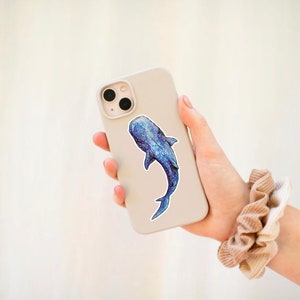 Op de afbeelding: Een witte telefoonhoes met een blauwe en witte walvishaai-sticker. De telefoonhoes wordt vastgehouden door een hand met een bruine scrunchie om de pols.