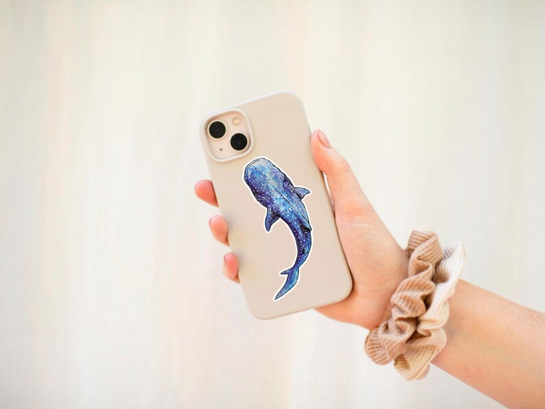 Op de afbeelding: Een witte telefoonhoes met een blauwe en witte walvishaai-sticker. De telefoonhoes wordt vastgehouden door een hand met een bruine scrunchie om de pols.