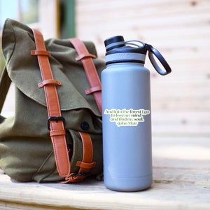 May include: A gray stainless steel water bottle with a black lid and handle. A sticker with the quote "And into the forest I go to lose my mind and find my soul. -John Muir" is on the bottle.
