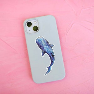 Op de afbeelding: Een lichtblauwe telefoonhoes met een walvishaai-sticker. De walvishaai is blauw en grijs met witte vlekken.