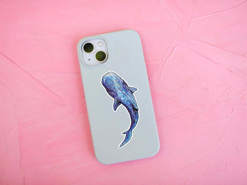 Op de afbeelding: Een lichtblauwe telefoonhoes met een walvishaai-sticker. De walvishaai is blauw en grijs met witte vlekken.