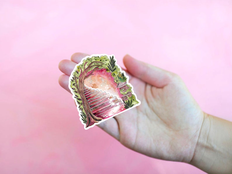 May include: A whimsical sticker featuring a watercolor illustration of a staircase leading into a glowing portal framed by a tree and foliage. The sticker has a white border and is held in a hand against a pink background.