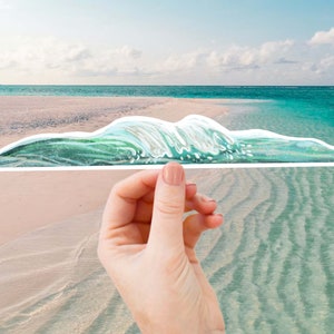 May include: A hand holds a sticker of a green and white watercolour wave, with a background of a beach and ocean.
