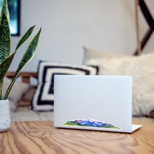 Pode incluir: Um computador port&aacute;til branco com um adesivo de aquarela de uma cordilheira na tampa. O adesivo apresenta tons de azul, roxo, verde e marrom. O laptop est&aacute; em cima de uma mesa de madeira.