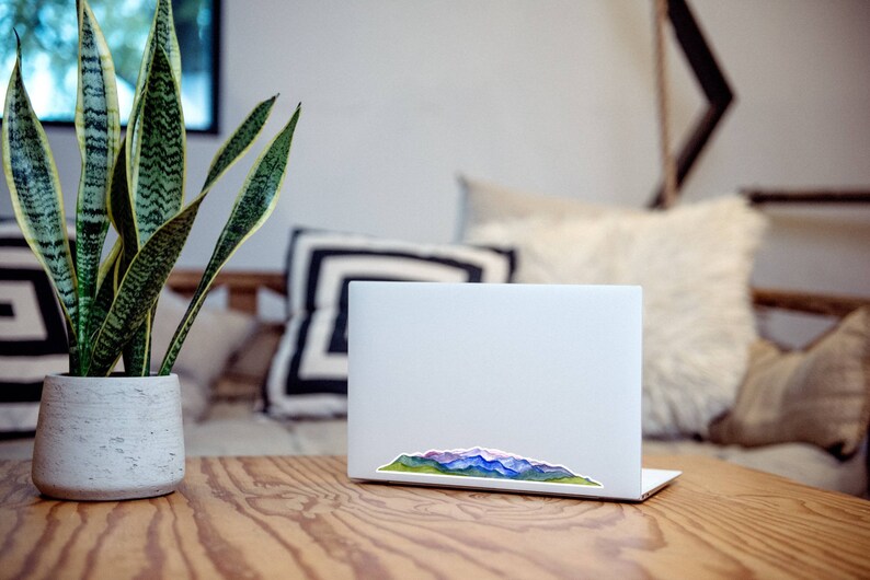 Pode incluir: Um computador port&aacute;til branco com um adesivo de aquarela de uma cordilheira na tampa. O adesivo apresenta tons de azul, roxo, verde e marrom. O laptop est&aacute; em cima de uma mesa de madeira.