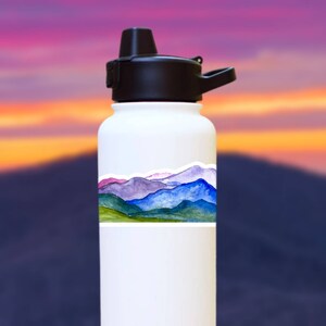 Pode incluir: Garrafa de &aacute;gua branca com um adesivo de uma cordilheira de montanhas em aquarela. O adesivo apresenta tons azuis, roxos, verdes e rosados.