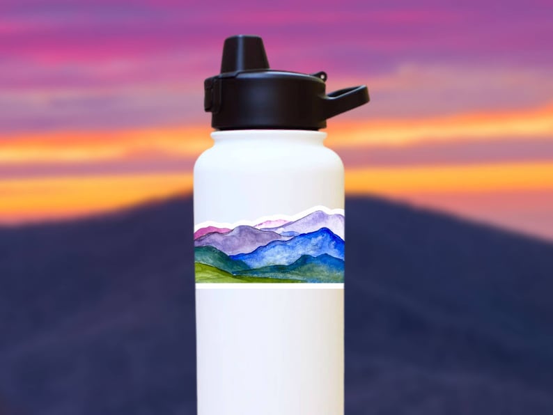 Pode incluir: Garrafa de &aacute;gua branca com um adesivo de uma cordilheira de montanhas em aquarela. O adesivo apresenta tons azuis, roxos, verdes e rosados.