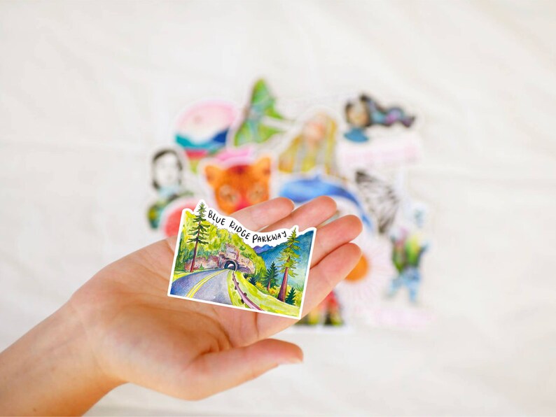 May include: A watercolor sticker featuring a scenic view of a road winding through a forest with a tunnel in the distance. The sticker has the text "Blue Ridge Parkway" printed on it.