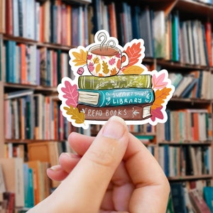 Op de afbeelding: Een sticker met een stapel boeken met de zinnen "Read Books" en "Support Your Library". Een bloemrijke theekop staat bovenop de boeken, omringd door herfstbladeren. De achtergrond is een wazige afbeelding van boekenplanken.