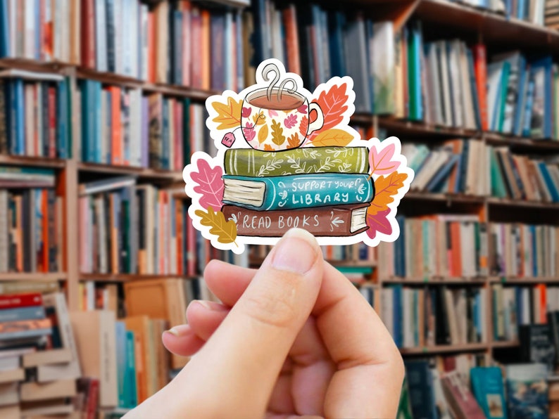 Op de afbeelding: Een sticker met een stapel boeken met de zinnen "Read Books" en "Support Your Library". Een bloemrijke theekop staat bovenop de boeken, omringd door herfstbladeren. De achtergrond is een wazige afbeelding van boekenplanken.