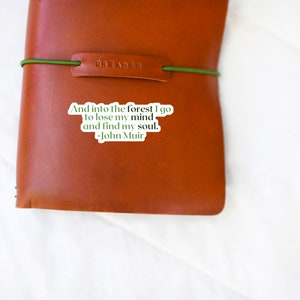 May include: A brown leather journal with a green elastic band and a sticker that reads "And into the forest I go to lose my mind and find my soul. -John Muir"
