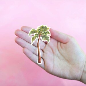 Puede incluir: Una mano sosteniendo un recorte de palmera, una ilustraci&oacute;n verde y marr&oacute;n sobre fondo blanco.