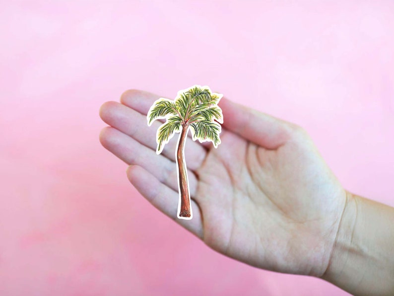Puede incluir: Una mano sosteniendo un recorte de palmera, una ilustraci&oacute;n verde y marr&oacute;n sobre fondo blanco.