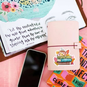 Op de afbeelding: Een roze leren notitieboek met een bordeauxrode elastische sluiting, een smartphone en een sticker van boeken en een mok. Een boek met de tekst "The Authenticity Project" is zichtbaar. Een journaalpagina heeft een citaat: "Laat de opwindende aantrekkingskracht van het volgende grote avontuur groter zijn dan de angst om het onbekende te betreden."