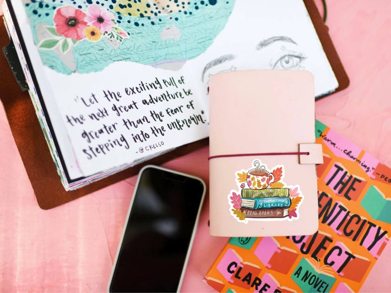Op de afbeelding: Een roze leren notitieboek met een bordeauxrode elastische sluiting, een smartphone en een sticker van boeken en een mok. Een boek met de tekst "The Authenticity Project" is zichtbaar. Een journaalpagina heeft een citaat: "Laat de opwindende aantrekkingskracht van het volgende grote avontuur groter zijn dan de angst om het onbekende te betreden."