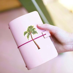 Puede incluir: Un diario de cuero rosa con una pegatina de palmera y un cierre de banda el&aacute;stica rosa.
