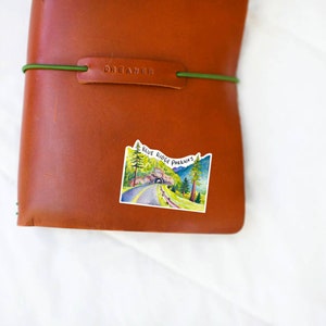 May include: A brown leather journal with a green elastic band and the word "DREAMER" embossed on the front. A sticker with a watercolor illustration of a road through a tunnel in the mountains is on the front of the journal. The sticker says "BLUE RIDGE PARKWAY".