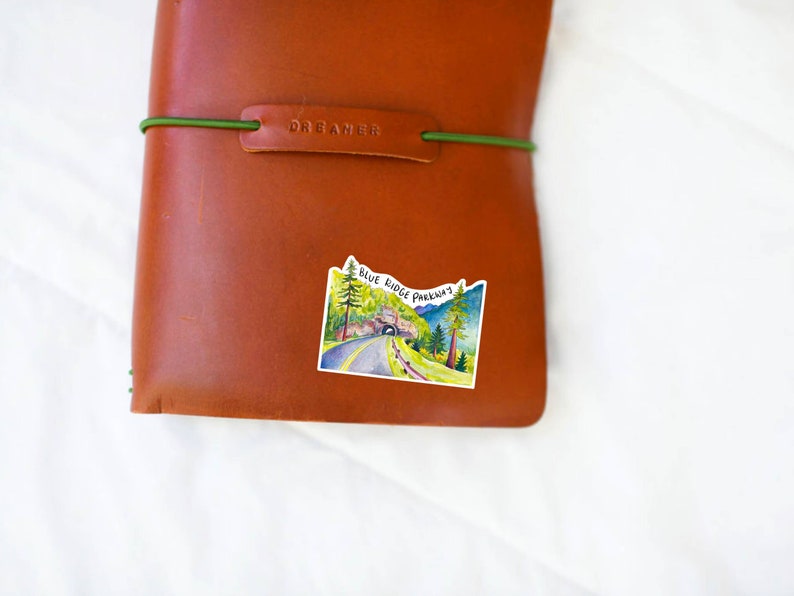 May include: A brown leather journal with a green elastic band and the word "DREAMER" embossed on the front. A sticker with a watercolor illustration of a road through a tunnel in the mountains is on the front of the journal. The sticker says "BLUE RIDGE PARKWAY".